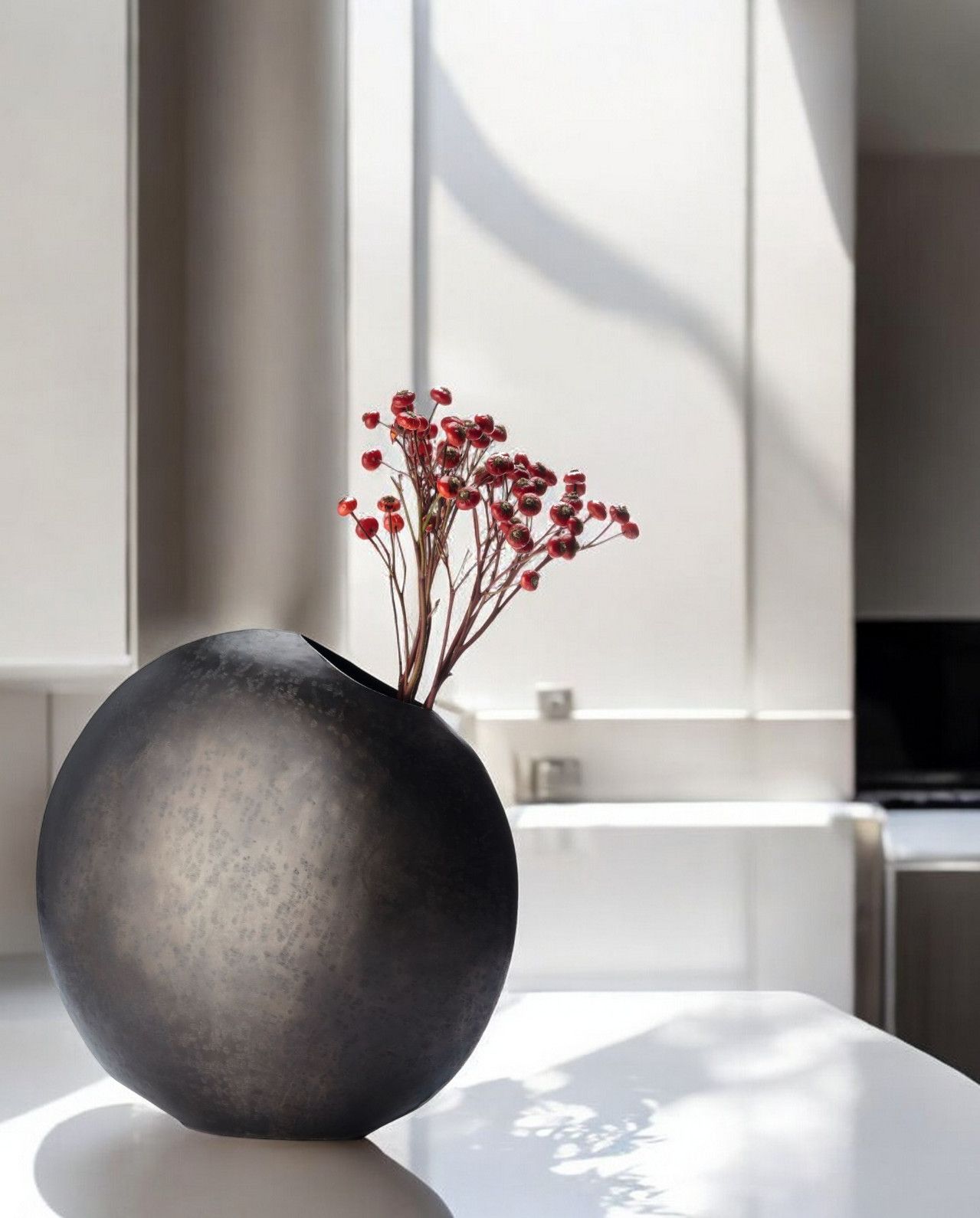 Monrovia Vase: Runde, dunkle Design-Vase mit roten Beeren-Zweigen als Dekoration auf einem weißen Tisch.