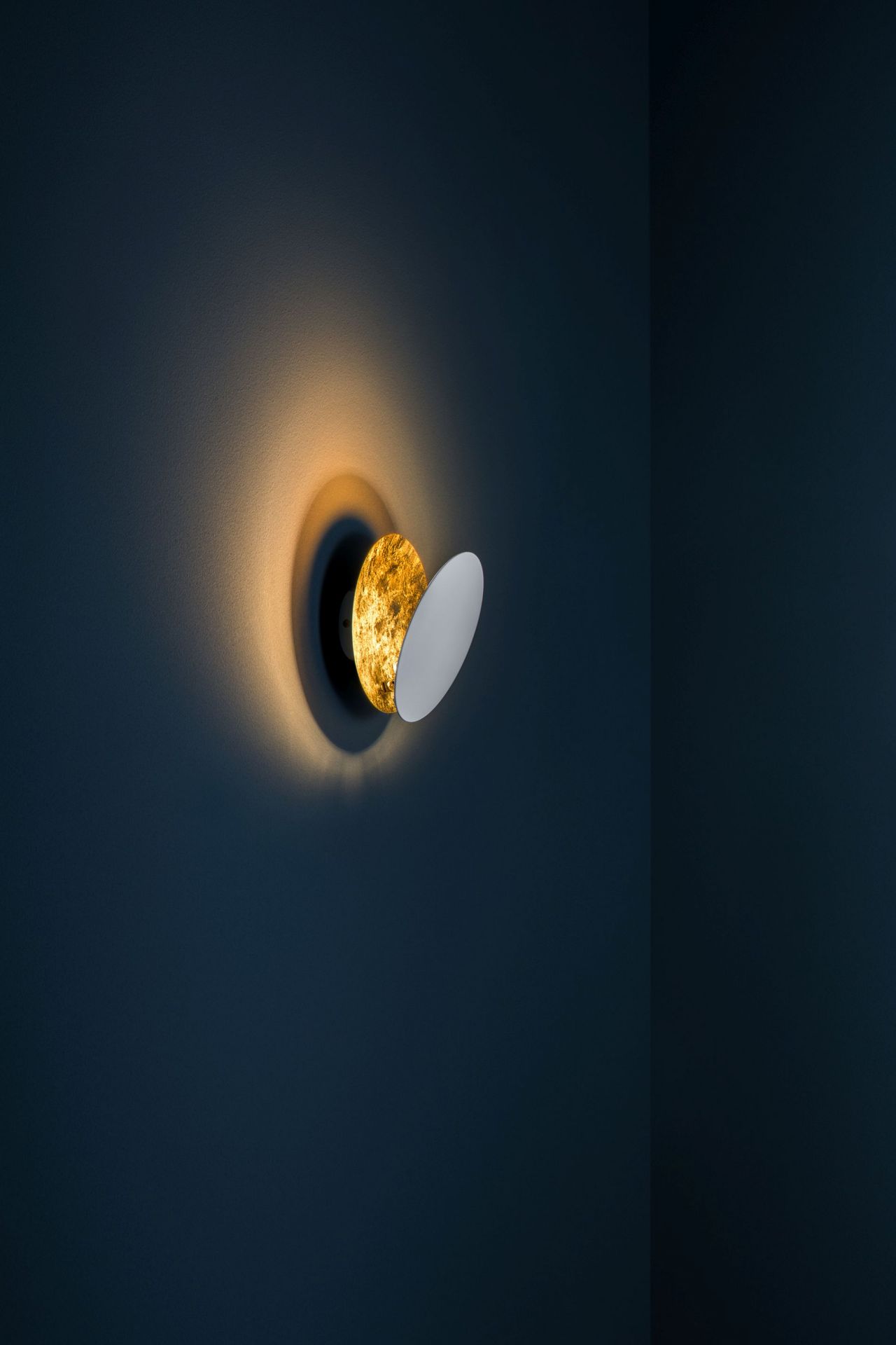 Lederam Wandleuchte von Catellani & Smith: Goldene Scheibe mit weißem Reflektor an dunkler Wand.