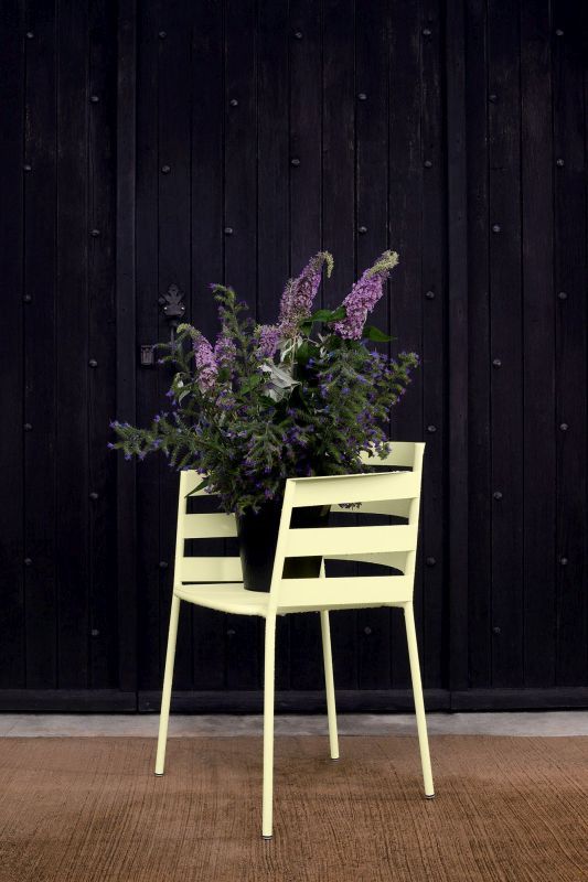 Heller Rythmic Sessel von Fermob für den Außenbereich mit lila Blumenstrauß vor dunkler Holzwand.