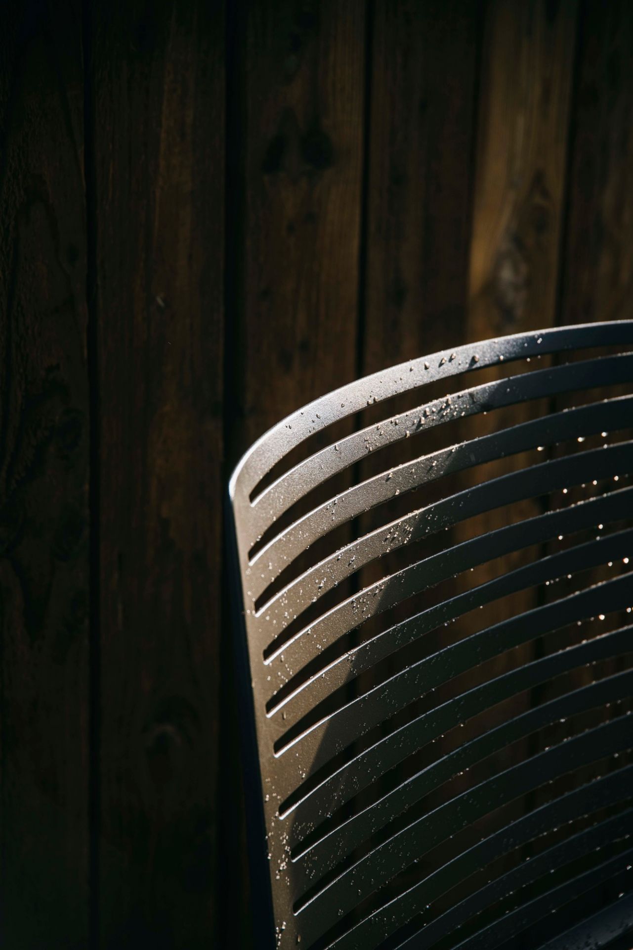 Detailaufnahme eines Zebra Outdoor Stuhls Fast aus Metall mit Wassertropfen vor dunkler Holzwand.