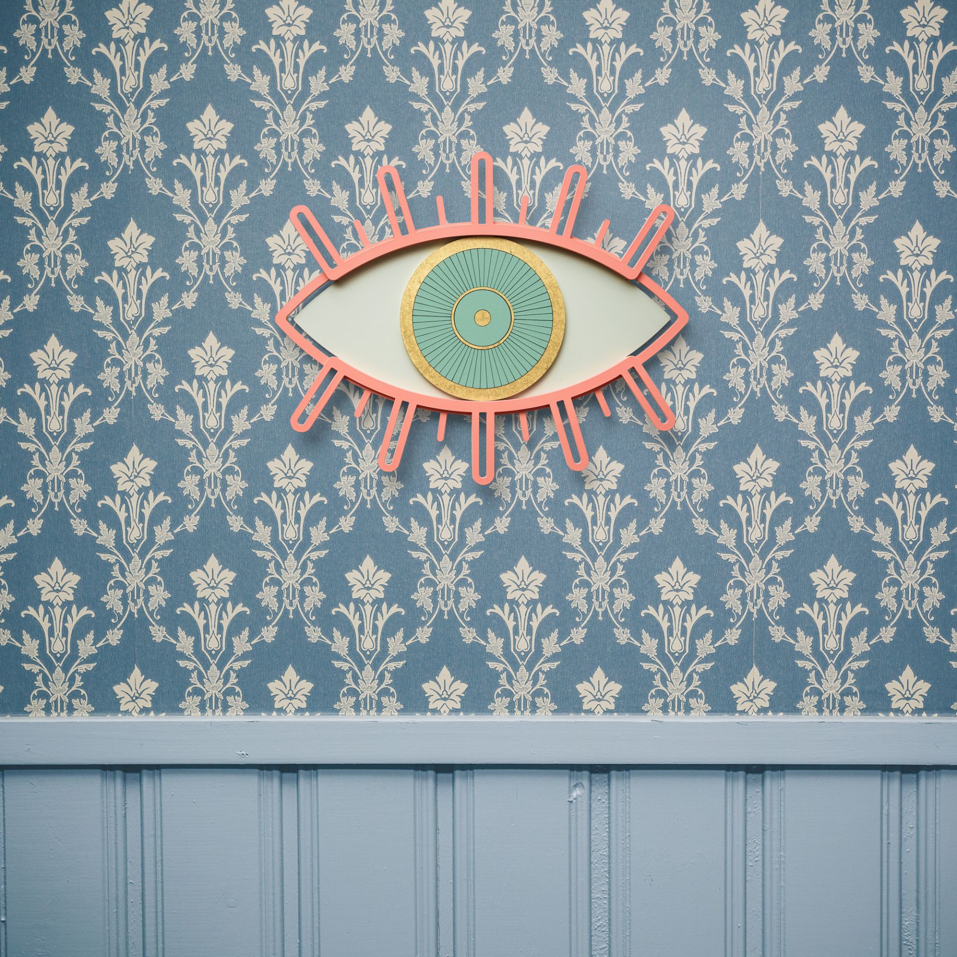 Eye 5 Wanddeko: Dekoratives Auge aus Holz in Rosa, Gold und Türkis an gemusterter Wand.
