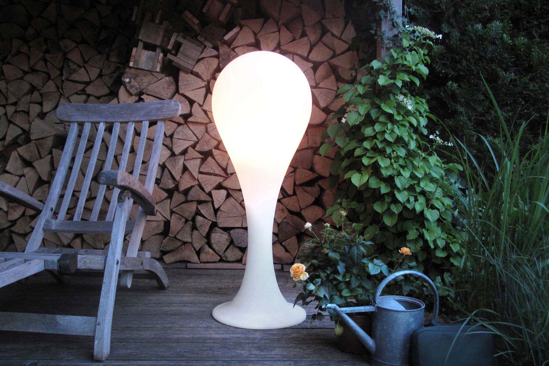 Liquid Light Drop Outdoor Standleuchte neben Holzstapel, Stuhl und Pflanzen im Garten.