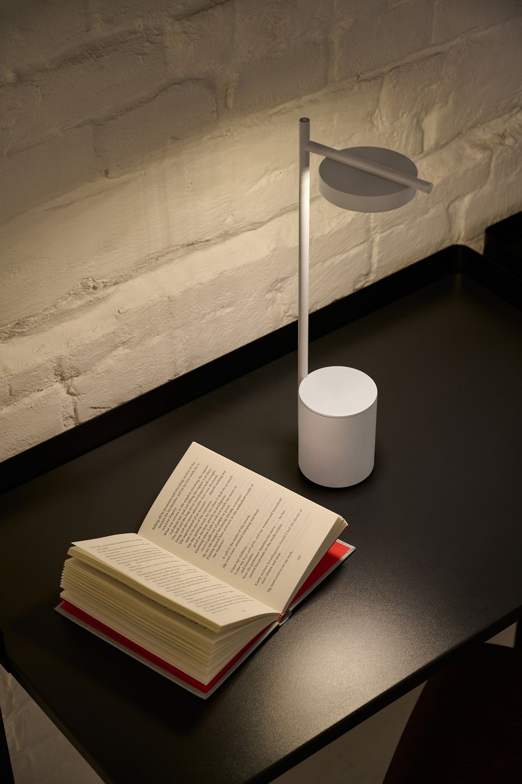 Weiße Igram Akku Tischleuchte von Grupa neben einem offenen Buch auf schwarzem Tisch.
