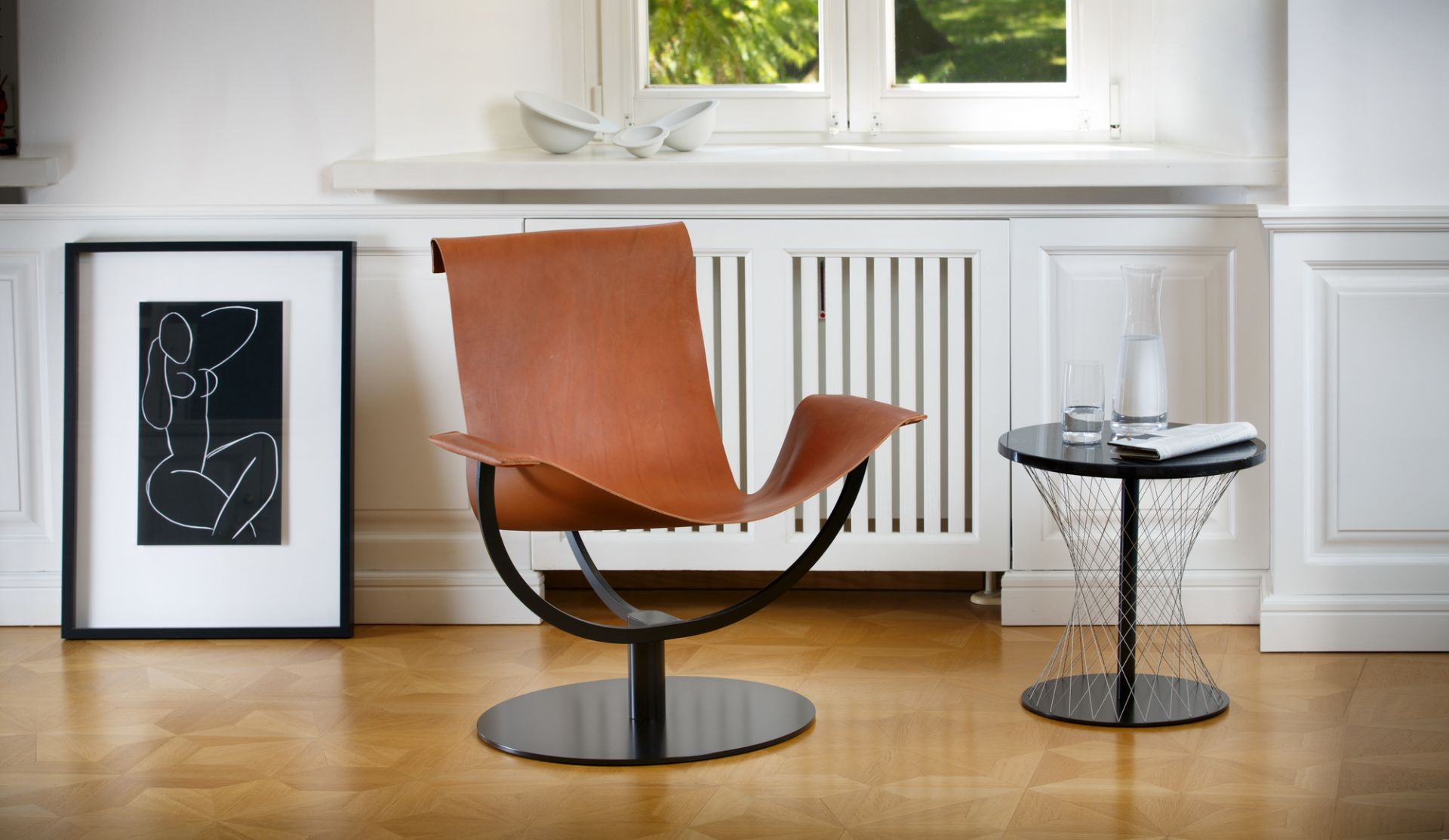 Schwarzer Network Beistelltisch Favius neben Design-Sessel mit Wasser und Glas auf Parkettboden.