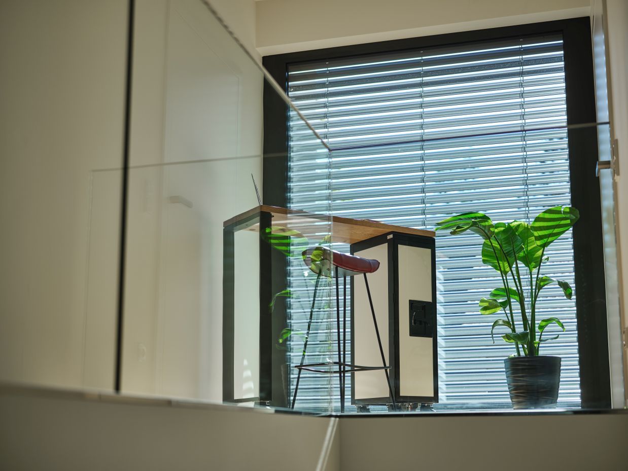 Schwarzer Janua Hightable Tisch mit Bordbar, rotem Barhocker und Zimmerpflanze vor einem Fenster mit Jalousien.