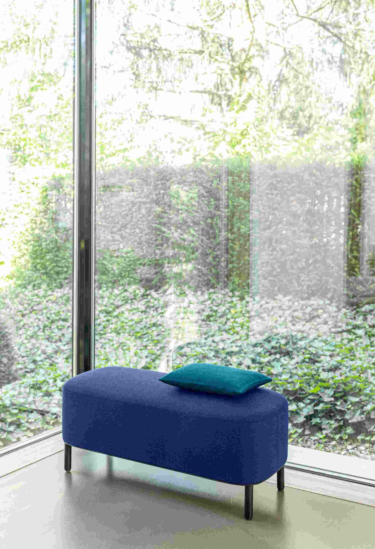 Blaue Amie Bank von Schönbuch mit türkisfarbenem Kissen vor großem Fenster. Moderne Sitzbank für Wohnzimmer.