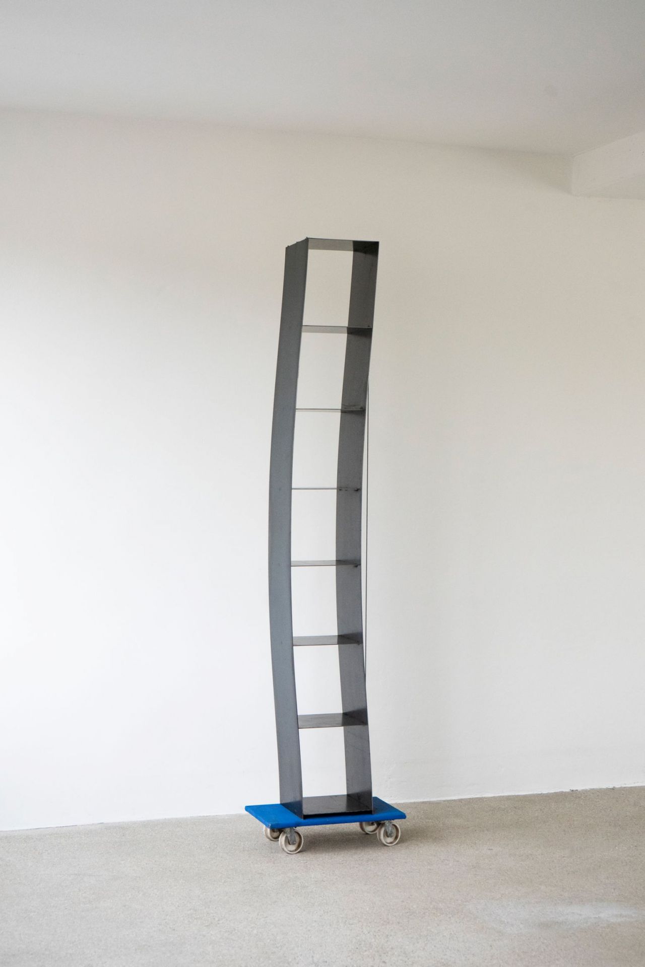 Gespanntes Regal von Moormann: Modernes, graues Bücherregal mit Glasböden auf blauem Rollbrett vor weißer Wand.