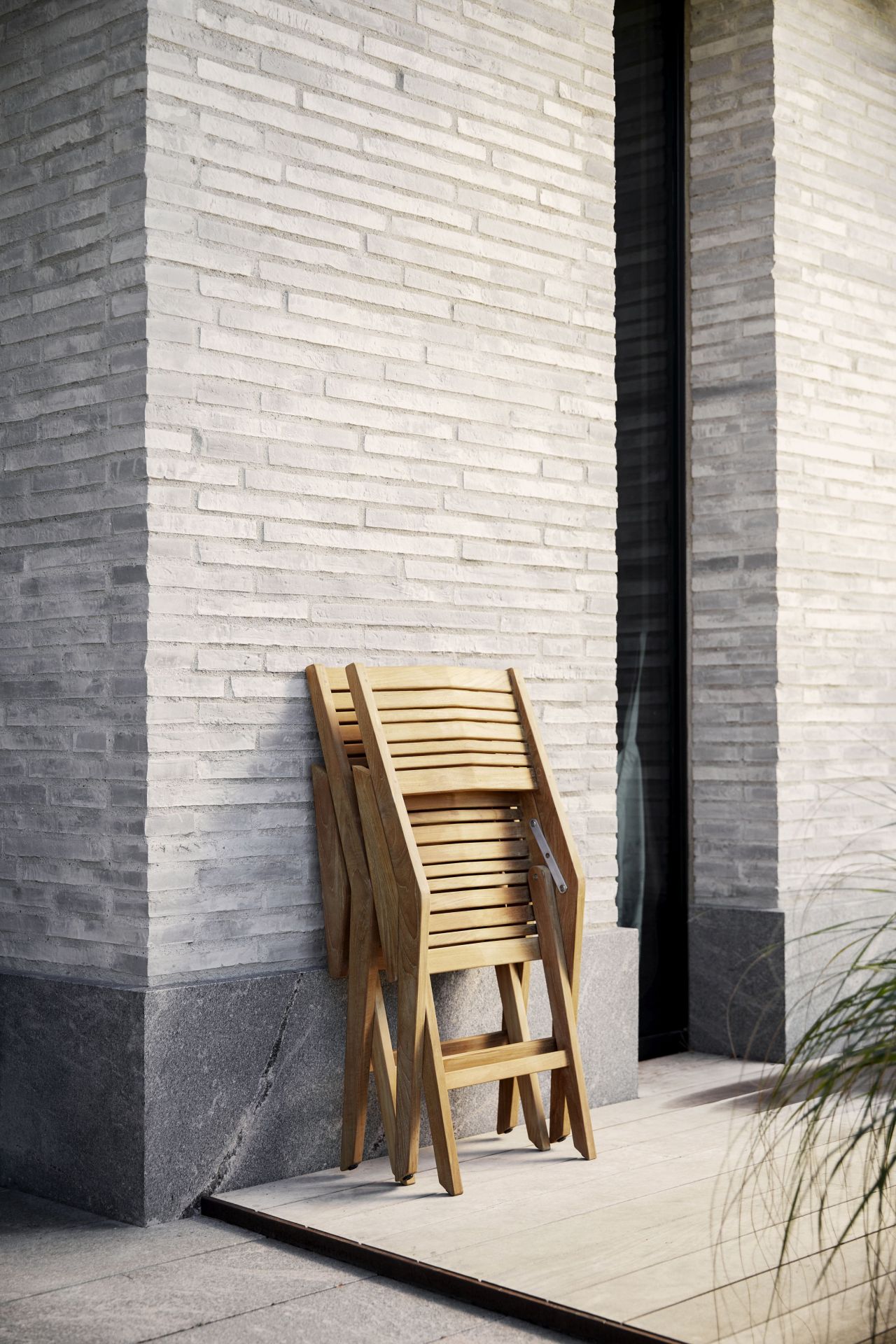 Zusammengeklappter Cane-Line Flip Armlehnstuhl aus Teakholz vor einer hellen Steinwand. Gartenmöbel für Balkon und Terrasse.