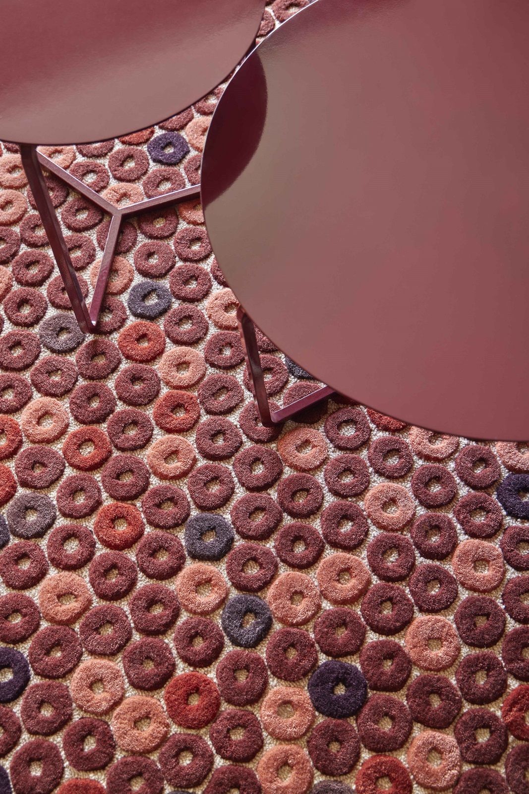 Detailaufnahme des Chimos Teppichs mit Donut-Muster in Rot- und Lilatönen, daneben zwei runde Tische.