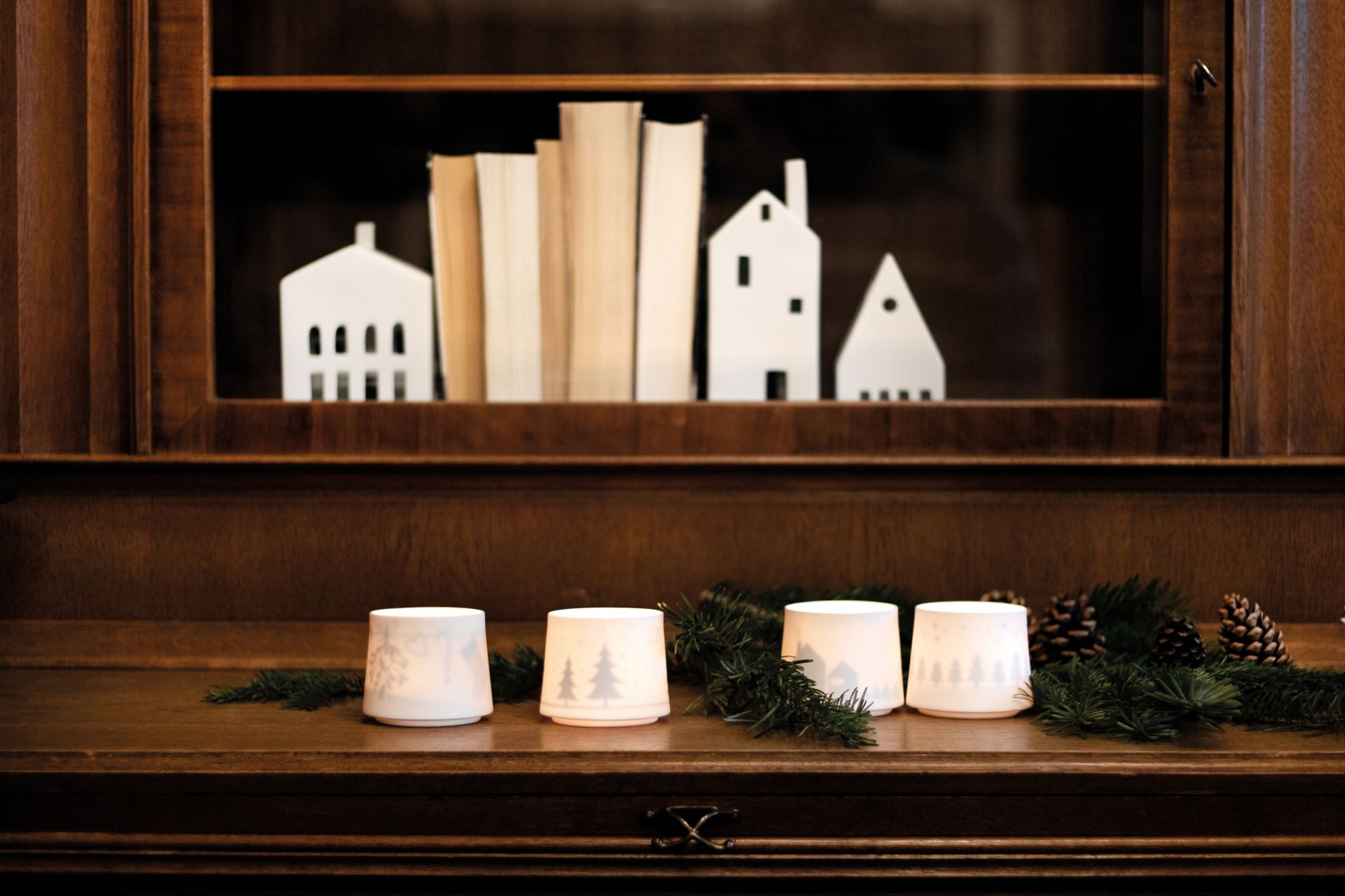 Dekoratives Zuhause Lichthaus mit Wintermotiven, Tannenzweigen und Zapfen auf einem dunklen Holzmöbelstück, stimmungsvolle Weihnachtsdekoration.