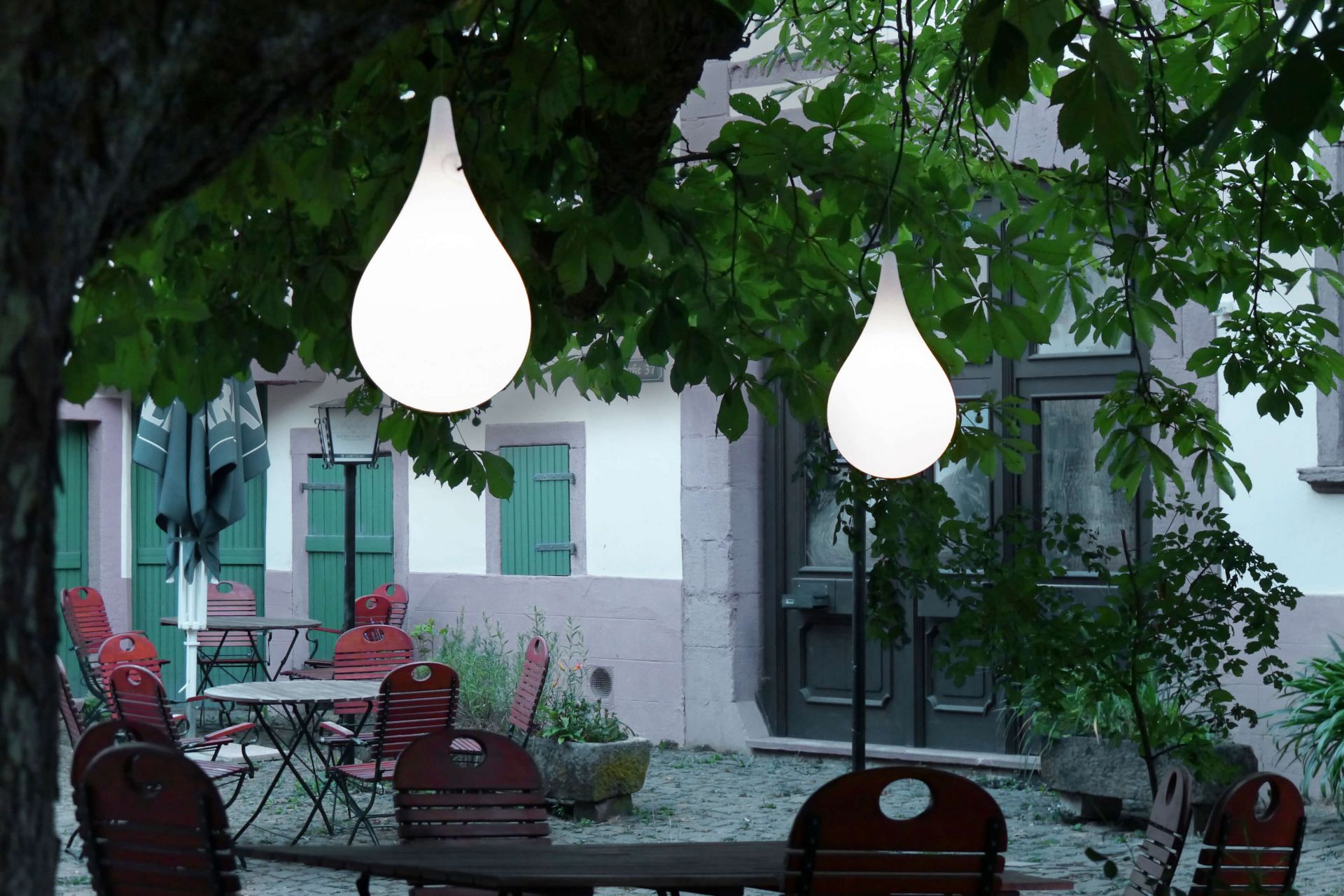 Zwei leuchtende Liquid Light Drop Pendelleuchten von Next im Außenbereich über Tischen und Stühlen.