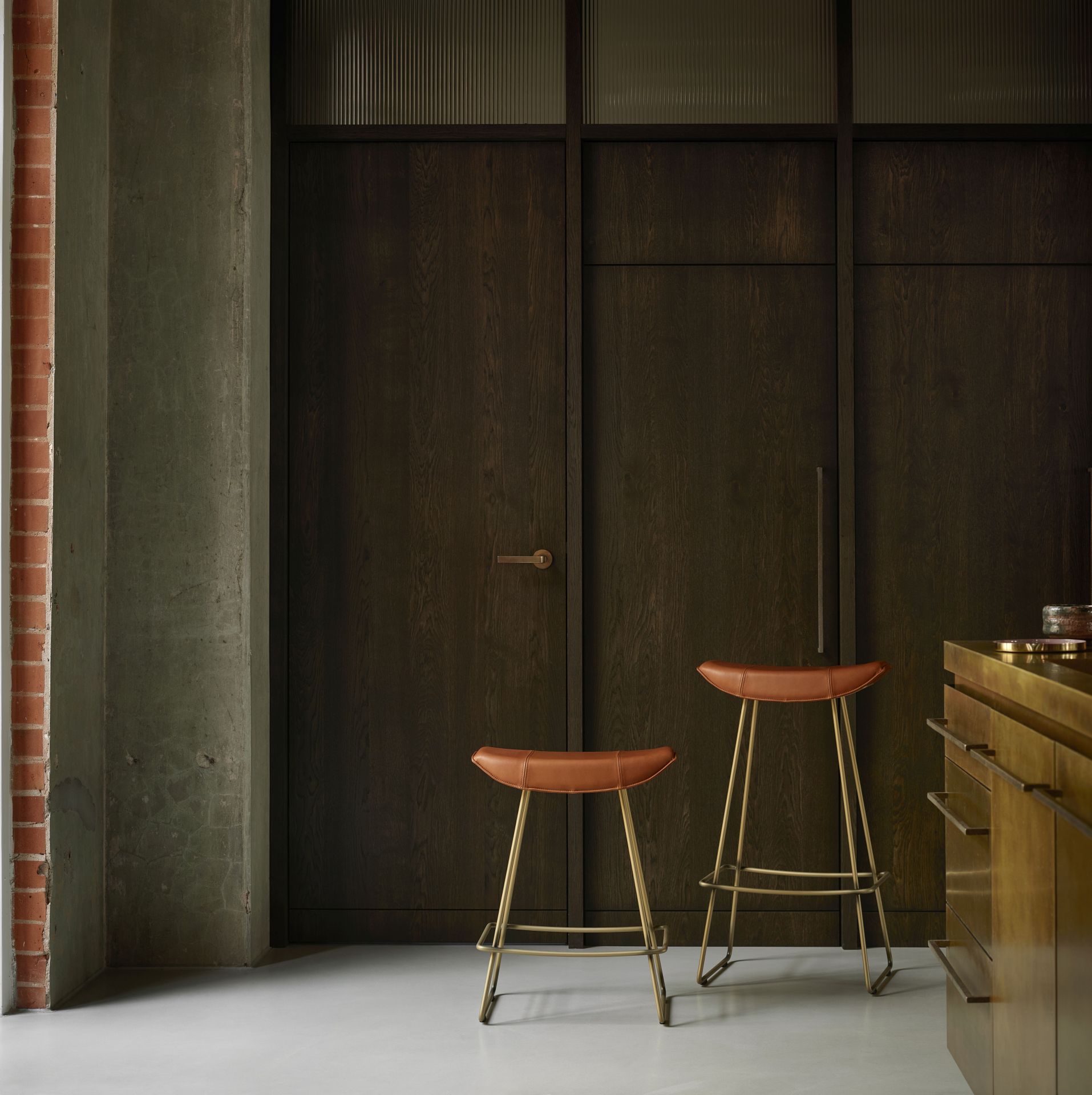 Zwei Kya Sitzhocker mit braunem Lederbezug und goldenem Gestell vor dunkler Holzwand.