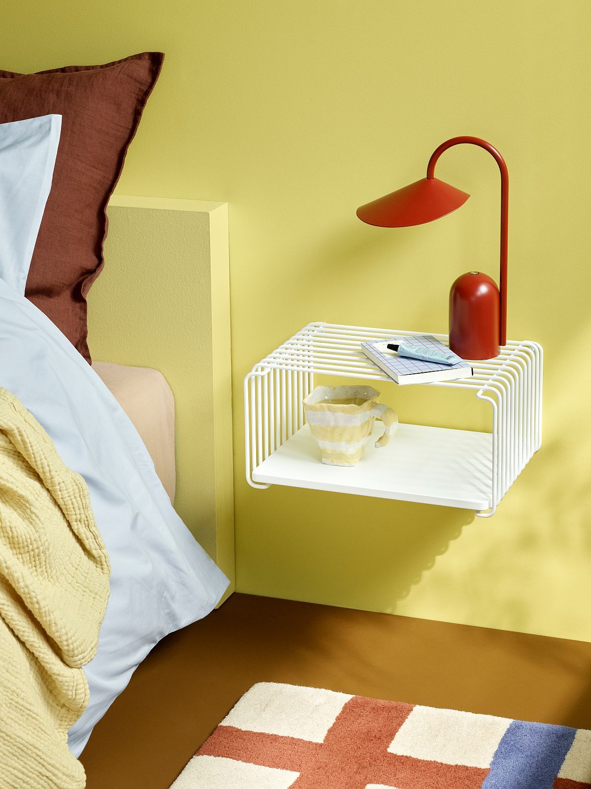 Weißes Panton Wire Regalsystem als Nachttisch mit Lampe, Tasse und Notizbuch vor gelber Wand.