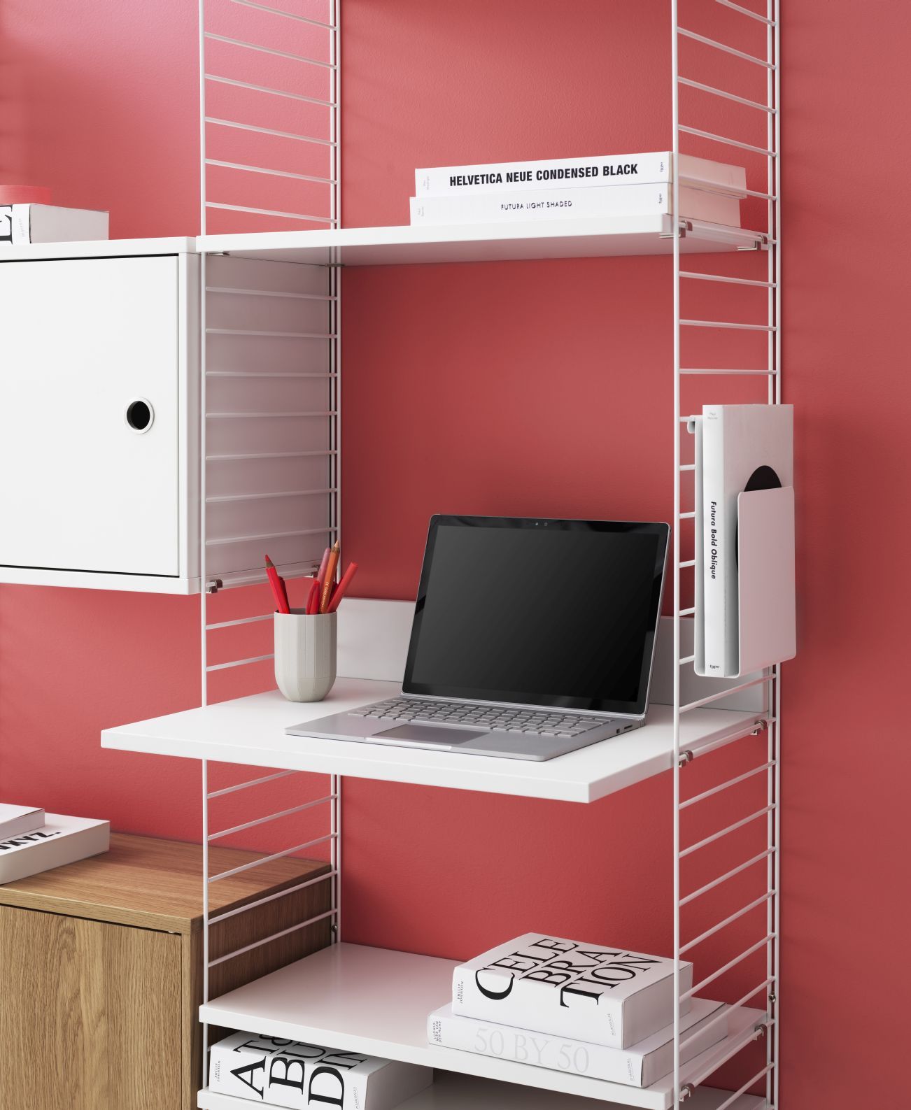 Beige String Regalsystem mit Schreibtisch, Laptop und Stauraum an roter Wand.