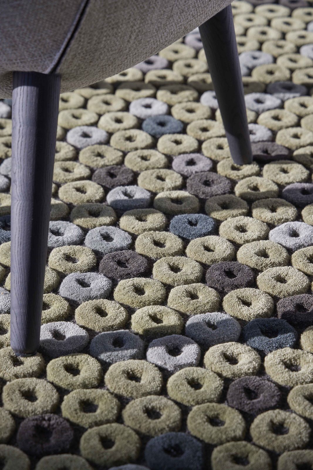Detailaufnahme des Chimos Rug Teppichs Gan mit Donut-Muster in Grau, Braun und Grün unter einem Stuhlbein.