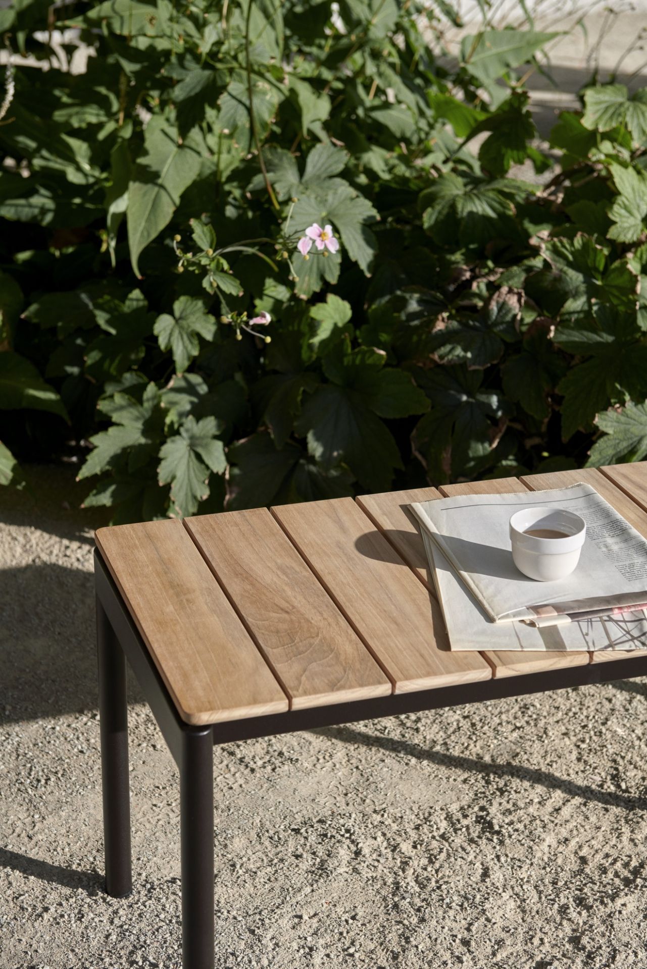 Nahaufnahme der AndTradition Ville AV27 Gartenbank aus Teakholz mit Zeitung und Kaffeetasse.