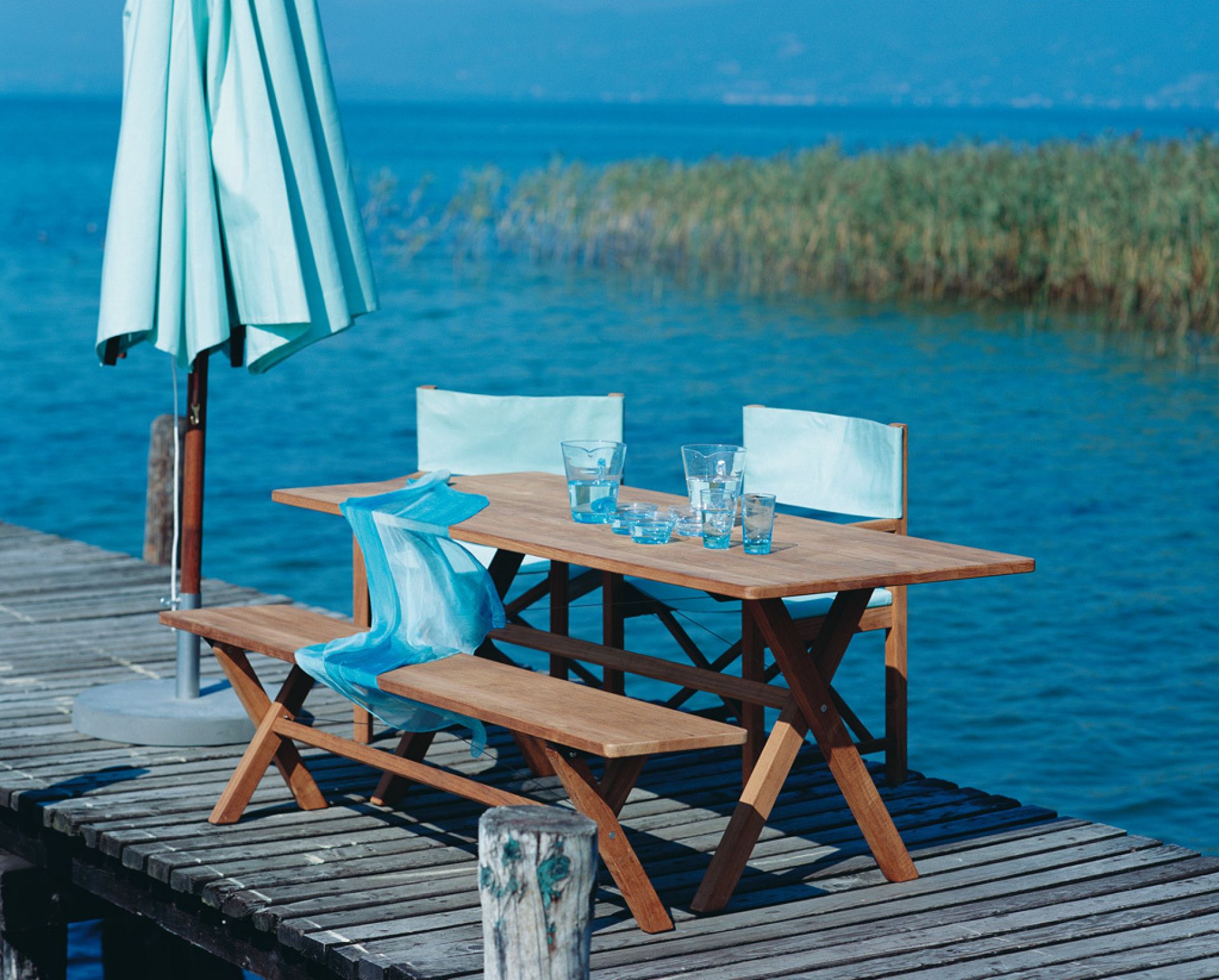 Cross Tisch klappbar von Weishäupl auf Holzsteg am See, mit Stühlen und Sonnenschirm.