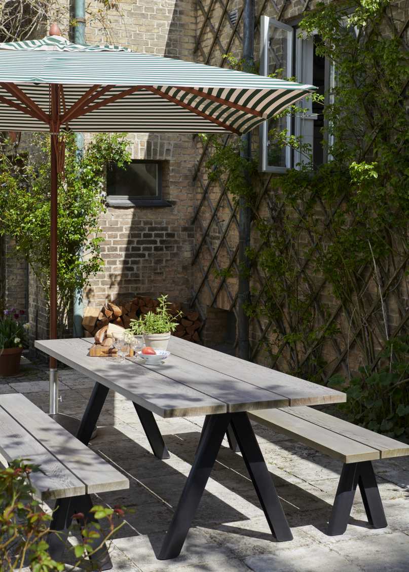 Outdoor-Szene mit Skagerak Overlap Table, Hunter Green, und Bänken unter einem Sonnenschirm.