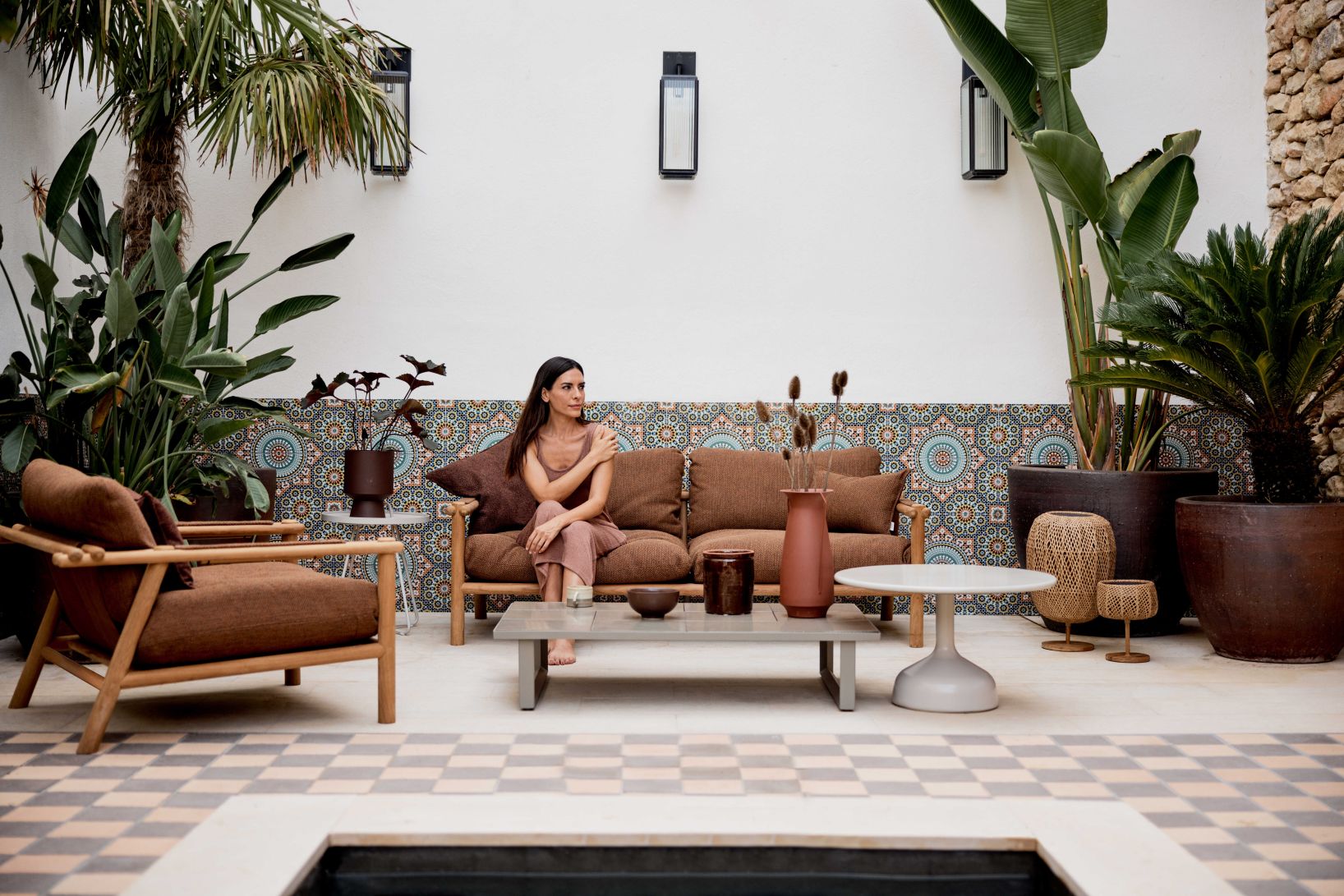 Frau sitzt auf Cane-Line Sticks Outdoor Loungesessel mit Kissen in einem begrünten Innenhof.