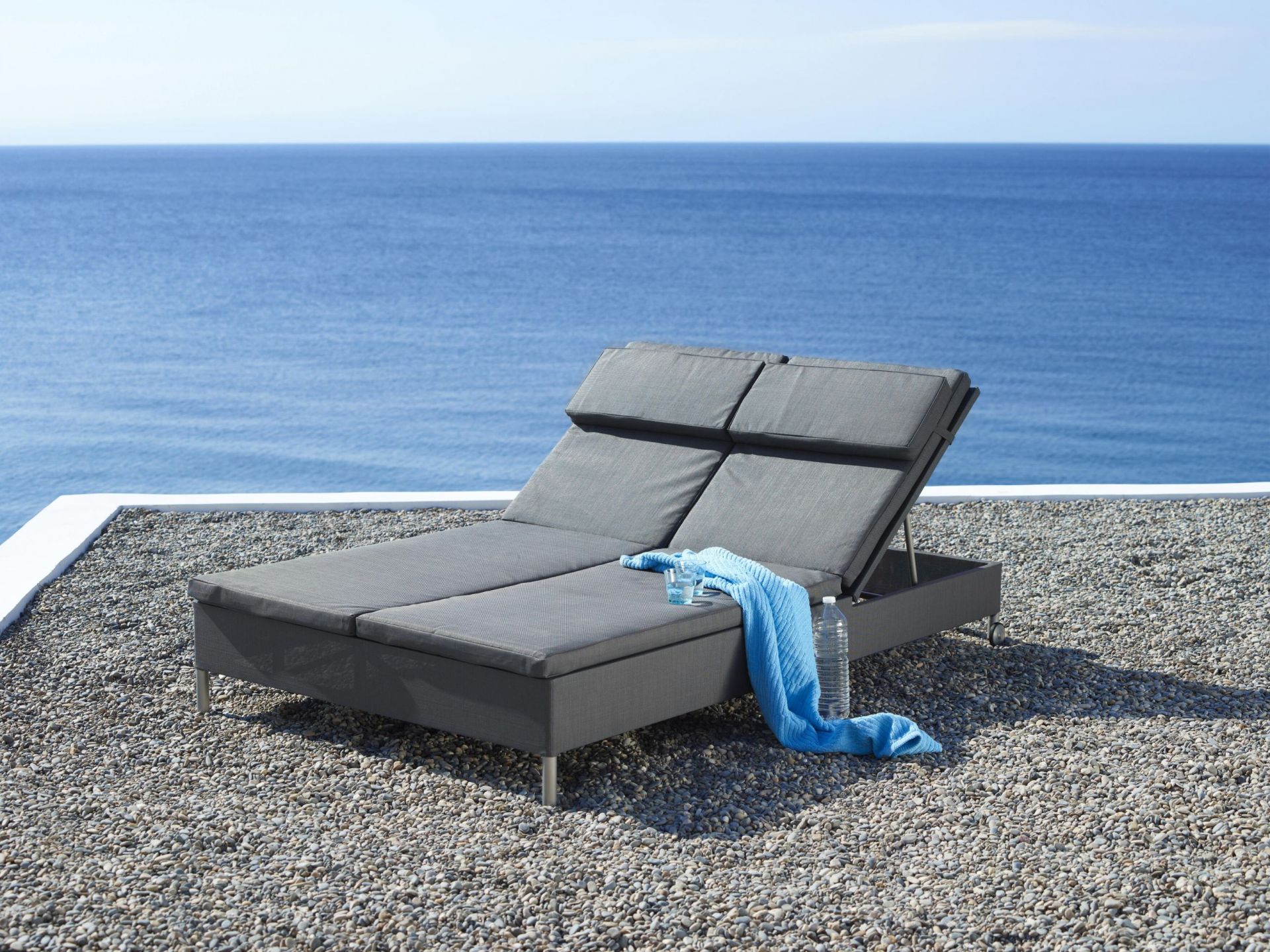 Graue Cane-Line Rest Sonnenliege für zwei Personen mit Handtuch und Getränken am Meer.
