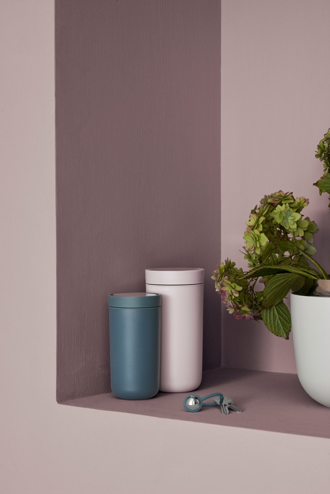 Zwei Stelton To Go Click Thermobecher in Blau und Weiß, daneben Schlüssel und Hortensie auf Regal.