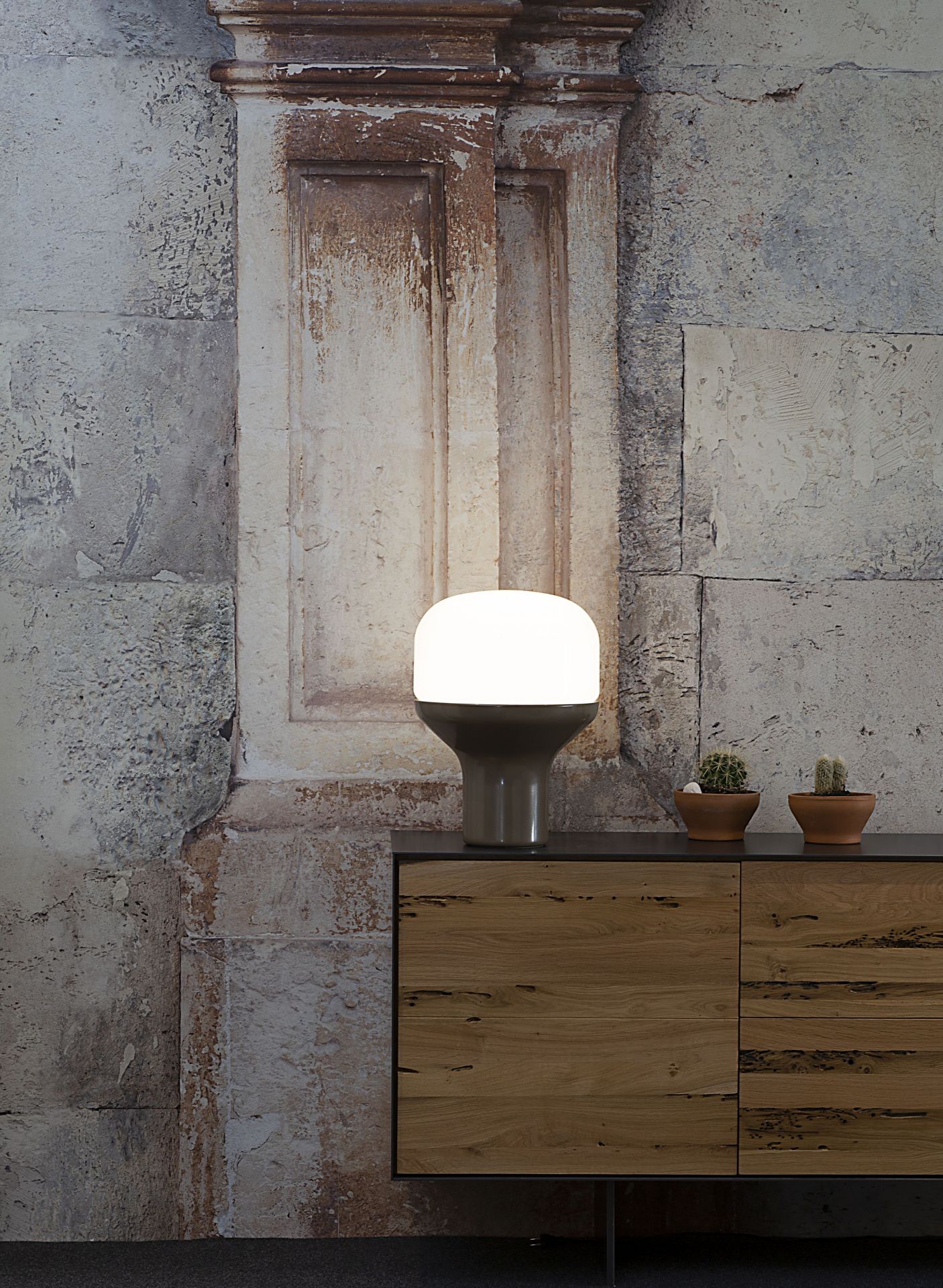 Delux Tischleuchte Sandgold von Martinelli Luce auf Holzkommode mit Kakteen vor Steinwand.