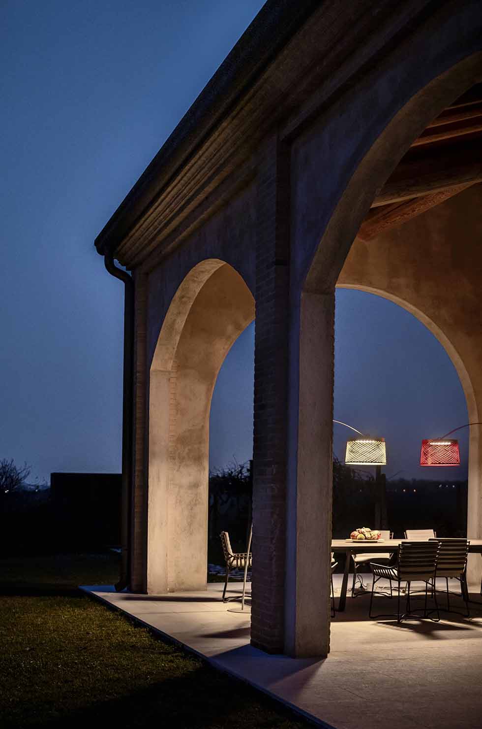 Twiggy Grid Stehleuchte kaminrot von Foscarini beleuchtet stimmungsvoll eine Terrasse mit Steinsäulen und Esstisch.