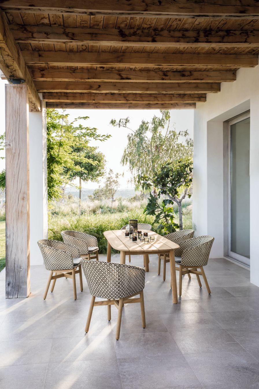 Mbrace Armlehnstuhl von Dedon mit Sitzkissen auf einer Terrasse mit Holztisch und Gartenblick.