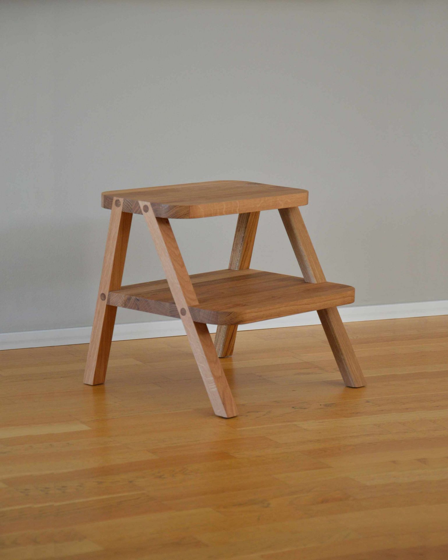 Oli Tritthocker von Jan Kurtz aus hellem Holz, zweistufig, auf Holzboden vor grauer Wand.