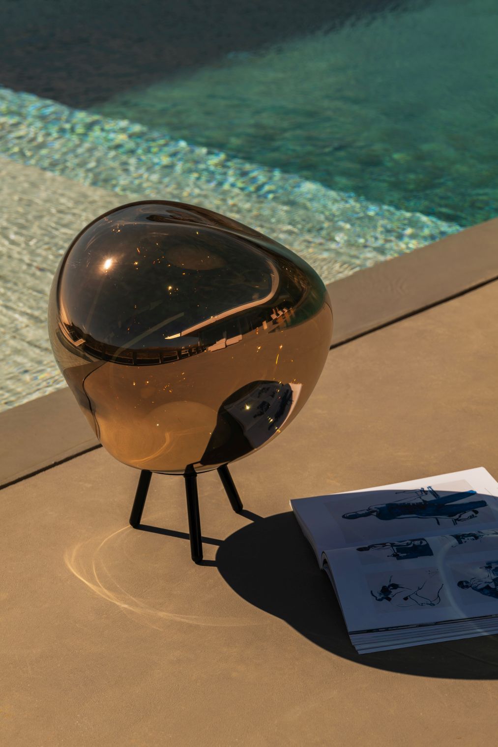 Kupferfarbene Tom Dixon Melt Leuchte auf drei Beinen neben einem Pool und Buch.