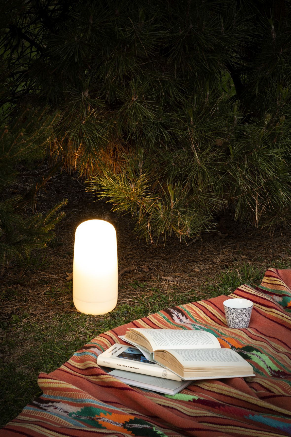 Artemide Gople Portable Akkuleuchte auf einer Decke im Freien, neben Büchern und einer Tasse.