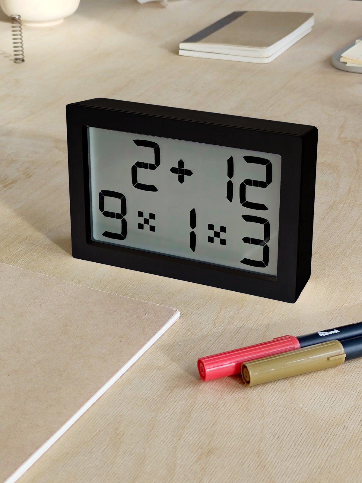 Schwarze Digitaluhr im Albert Clock Design auf einem Schreibtisch mit Stiften und Notizbüchern.
