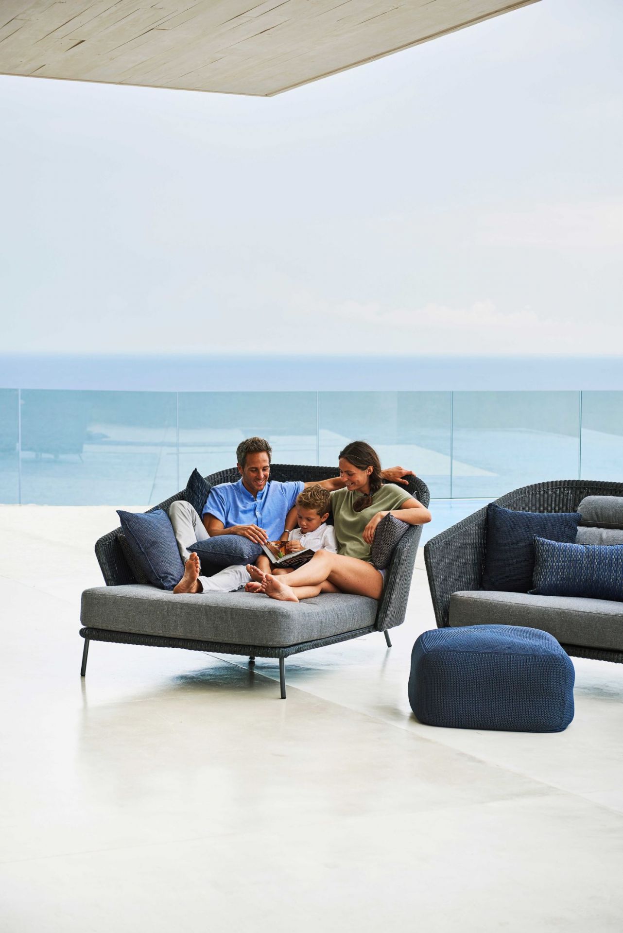 Familie entspannt auf Cane-Line Mega Outdoor Daybed mit grauer Polsterung und Blick aufs Meer.