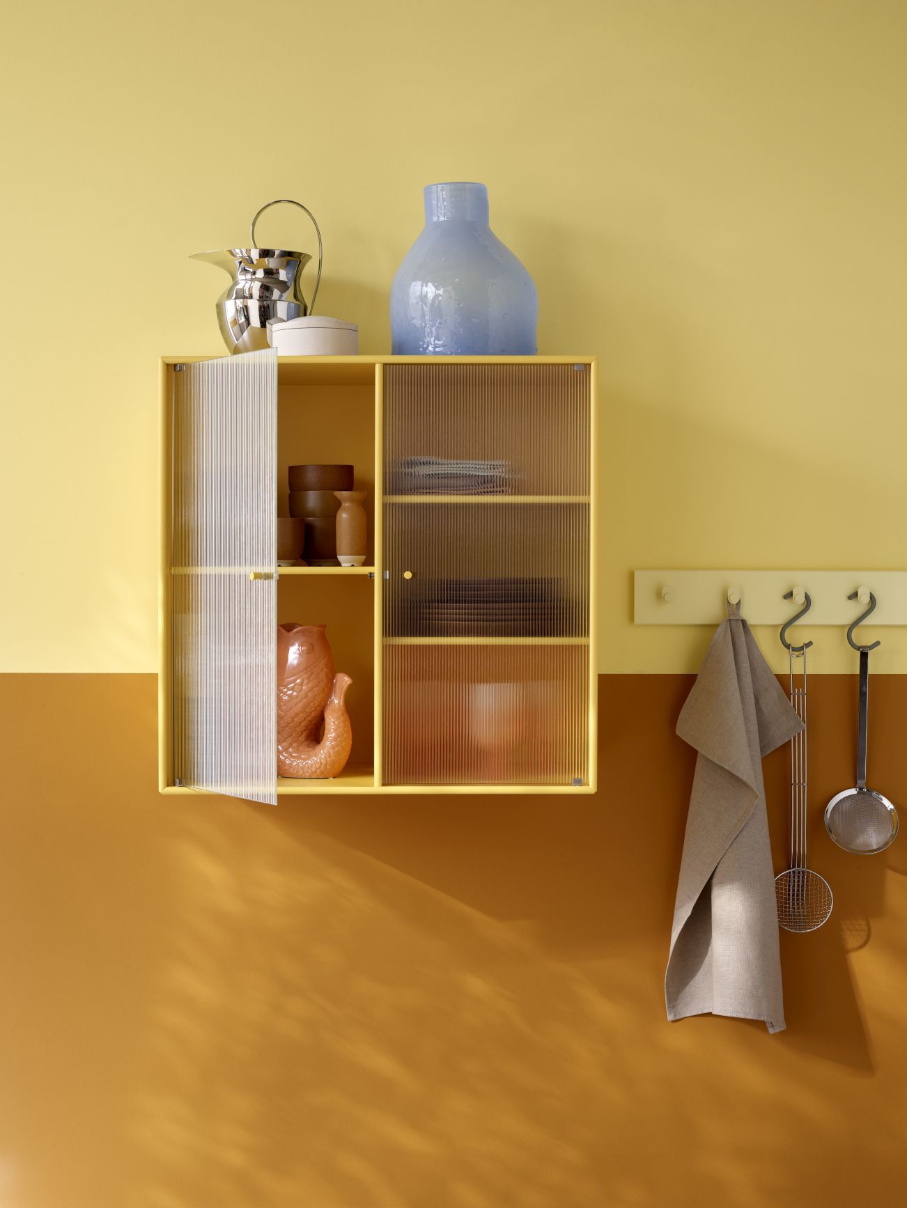 Gelber Ripple II Wandschrank mit geriffelten Glastüren, gefüllt mit Dekoration, an zweifarbiger Wand.