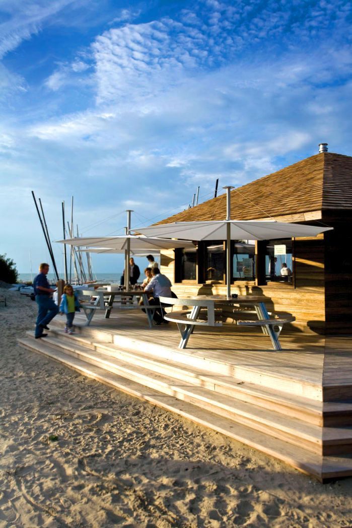 Extremis Gargantua Picknicktisch auf Holzterrasse am Strand mit Personen und Sonnenschirmen. Outdoor-Möbel für Gastronomie.