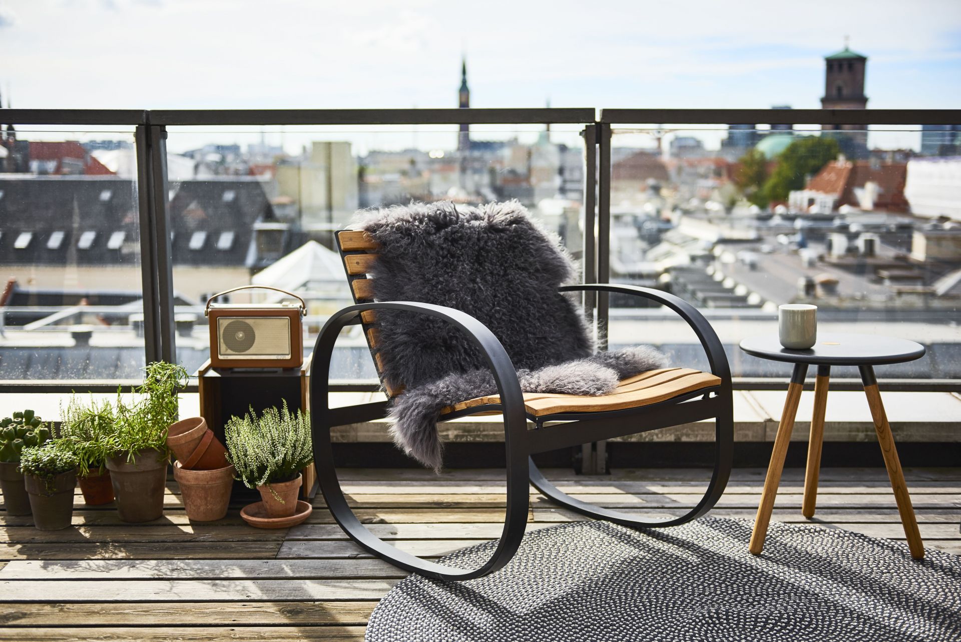 Parc Schaukelstuhl von Cane-Line auf einer Dachterrasse mit Schaffell und Beistelltisch. Outdoor Möbel für entspannte Momente.