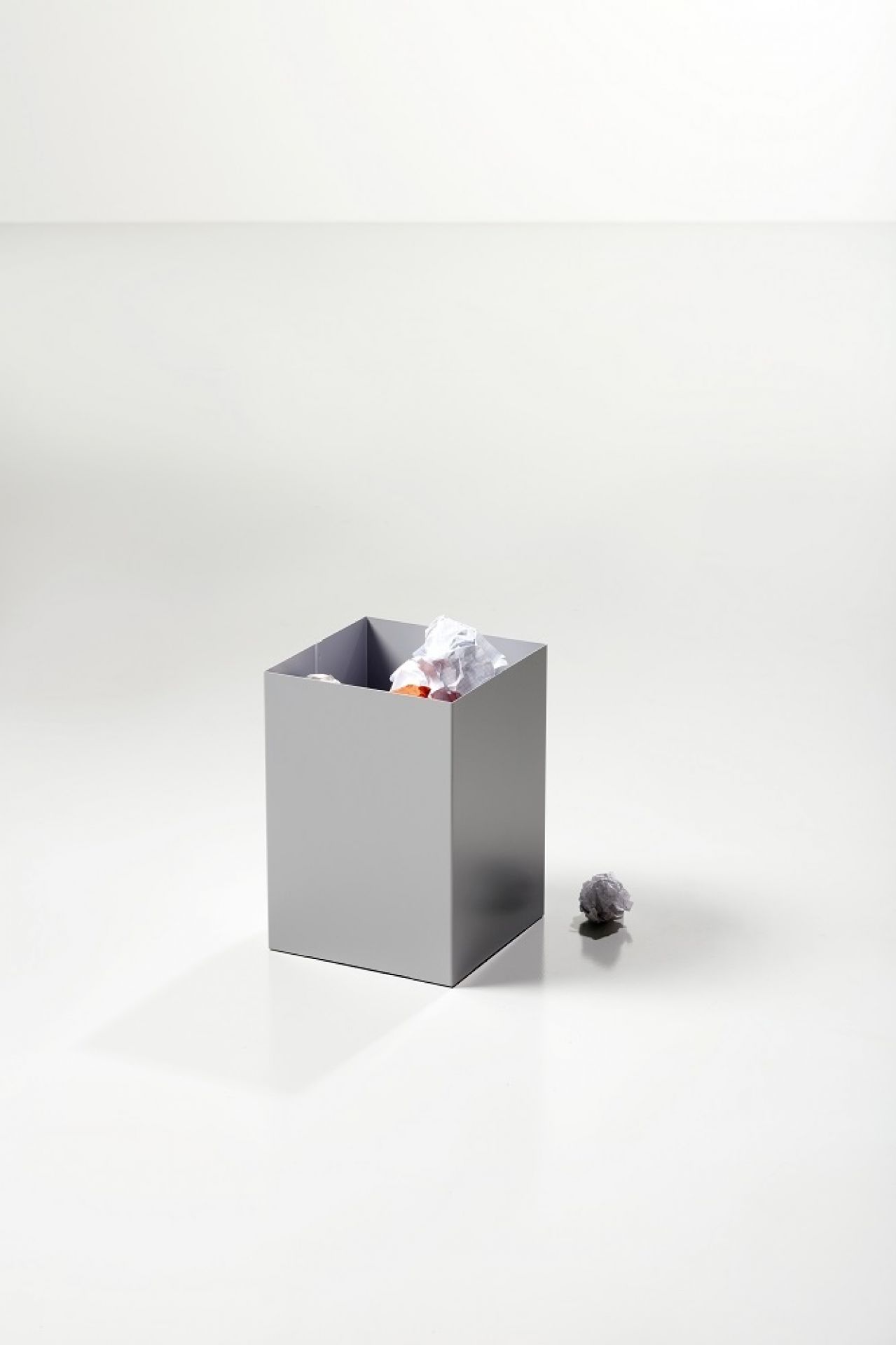 Grauer, quadratischer Cascando Papierkorb mit Papierknäueln, Büromüll und einem zerknüllten Papierball daneben.