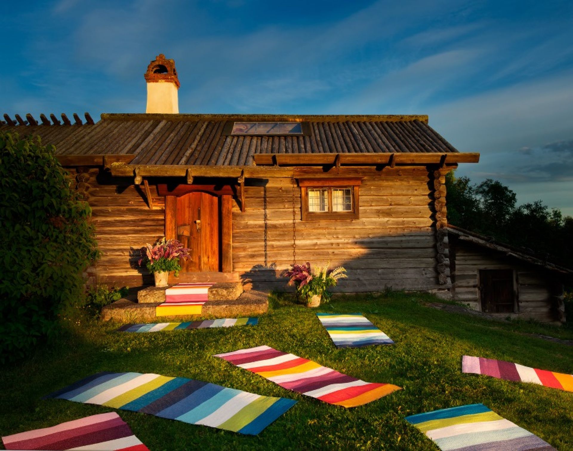 Mehrere gestreifte Molly Kunststoffteppiche von Pappelina im Außenbereich vor einem Holzhaus.