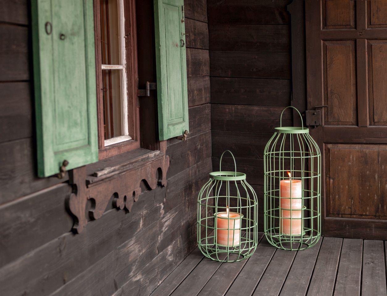 Zwei grüne Weishäupl Outdoor Laternen mit brennenden Kerzen vor einer Holzwand und Fenster.