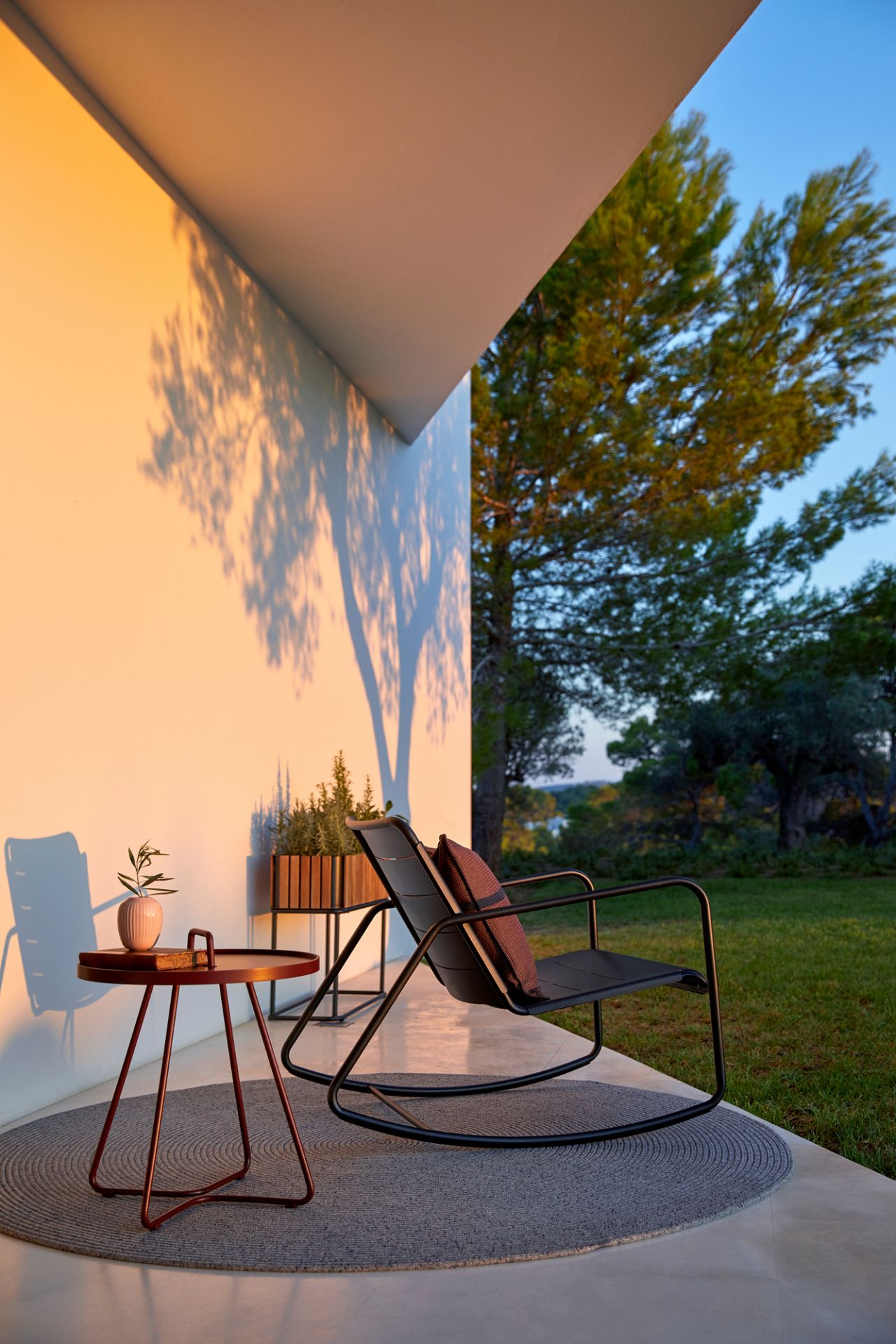 Cane-Line Schaukelstuhl für den Außenbereich mit Kissen auf Terrasse mit Beistelltisch und Pflanze.