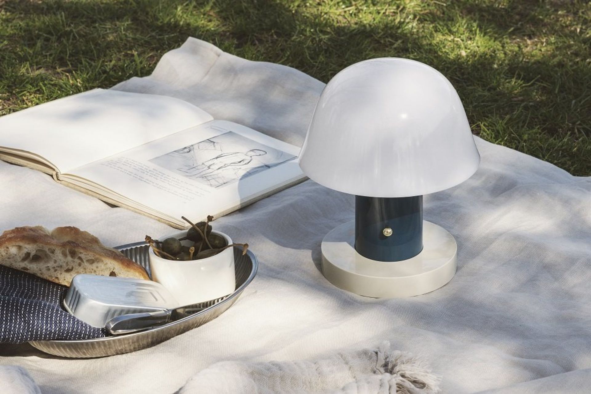 Setago Akkuleuchte Twilight & Sand von AndTradition auf Picknickdecke mit Brot, Oliven und Buch.