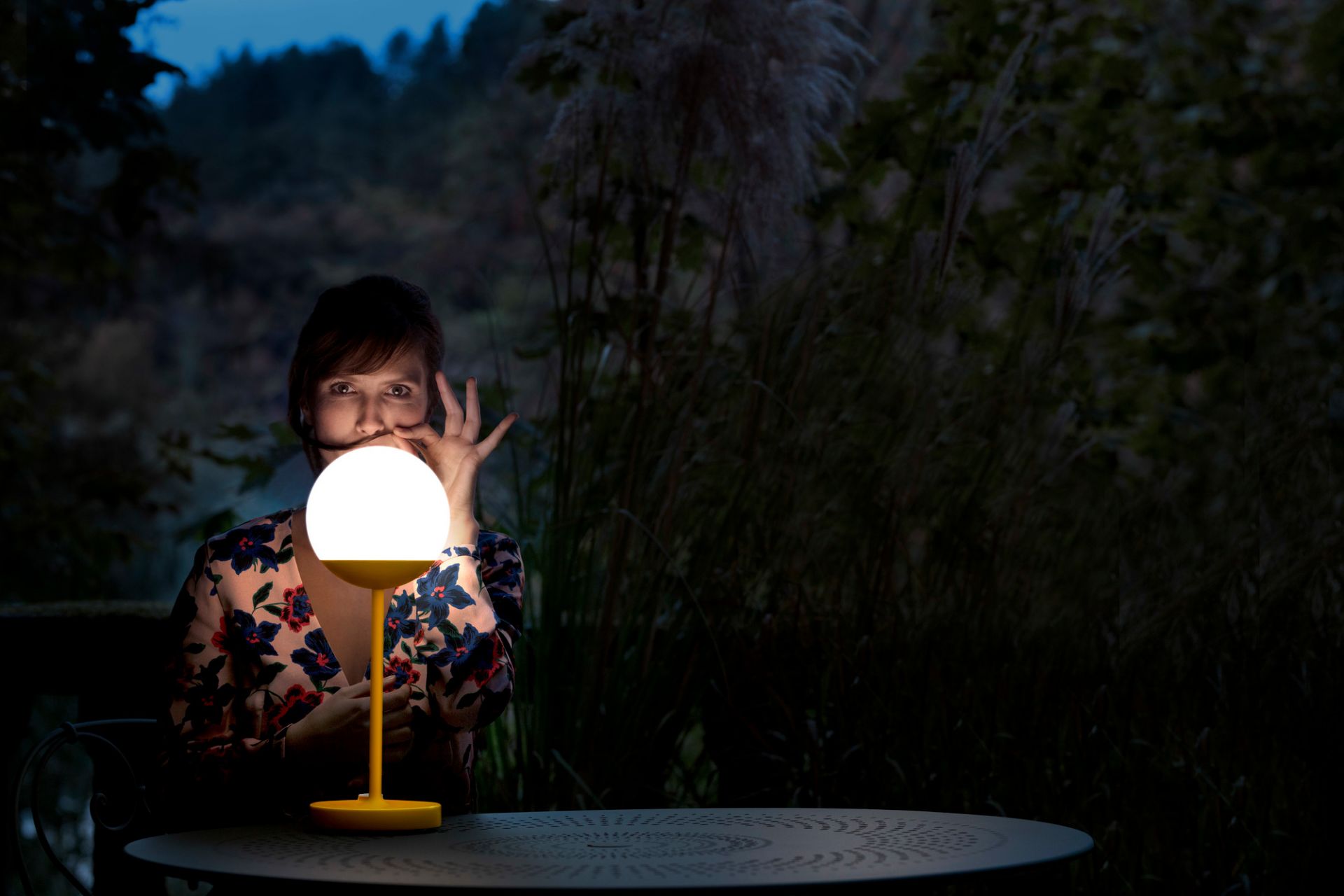 Mooon Tischleuchte von Fermob, anthrazitfarbener Tisch, Frau mit Blumenmuster-Bluse im Garten. Dekorative Leuchte für stimmungsvolle Beleuchtung.