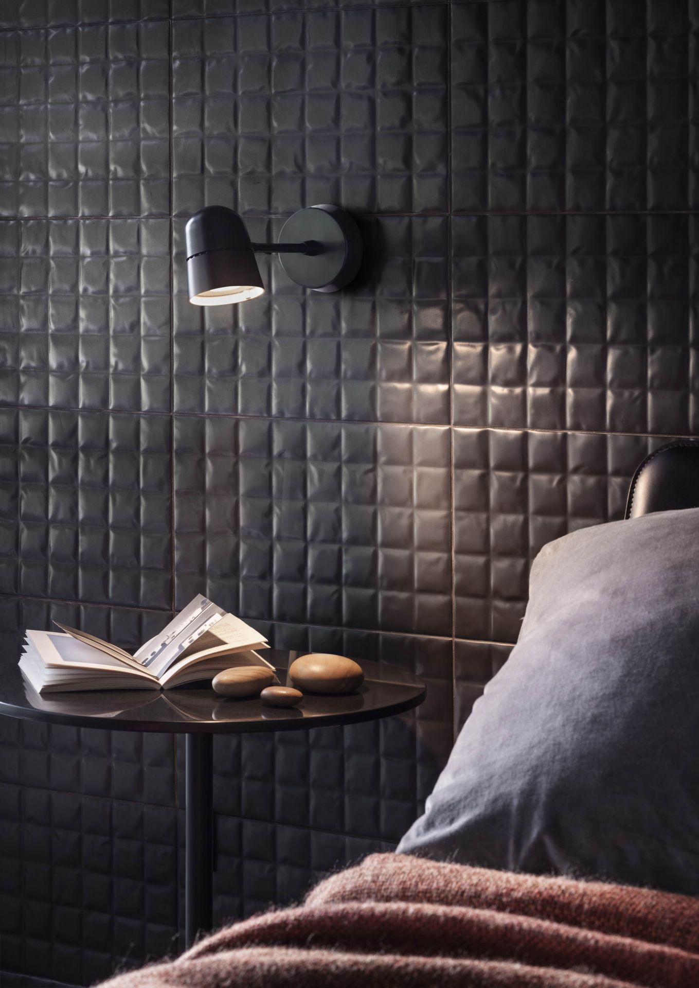 Schwarze Luceplan Spot Wandleuchte neben Bett mit Buch und Deko-Steinen auf Nachttisch.