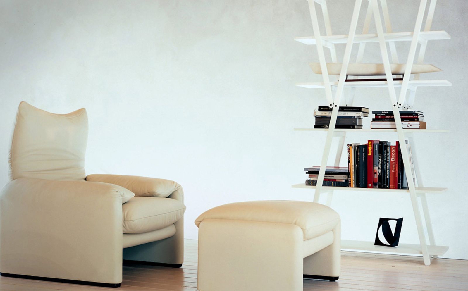 Beiger 675 Maralunga Hocker von Cassina neben Sessel und weißem Bücherregal für modernes Wohnzimmer.