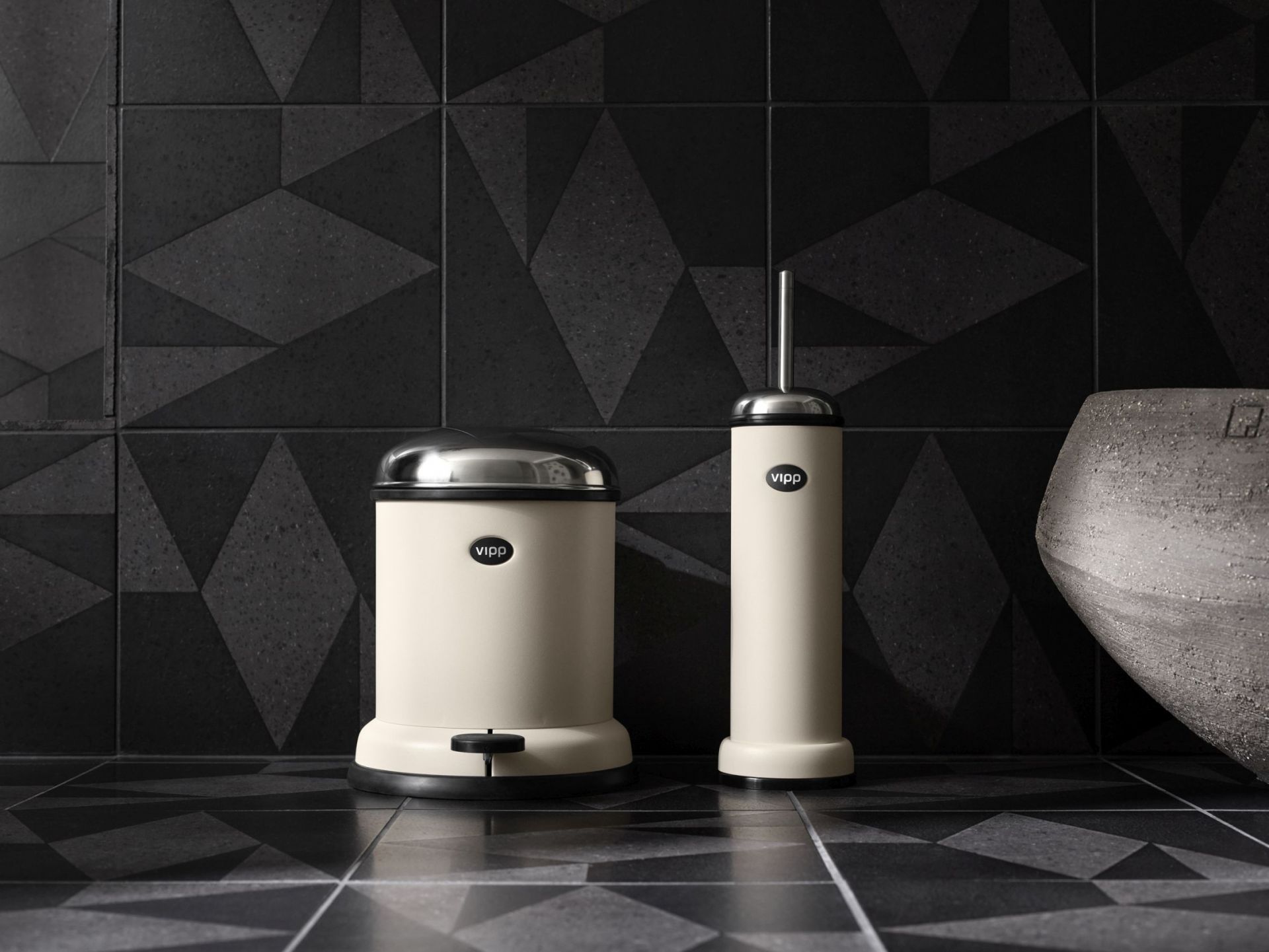 Beige Vipp WC-Bürste und Treteimer aus Edelstahl vor dunkler, geometrischer Wandfliese für moderne Badezimmerausstattung.
