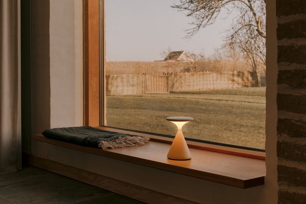 Graue Salt Akkuleuchte als Tischleuchte auf Fensterbank mit Decke, warmes Licht, stimmungsvolle Beleuchtung.