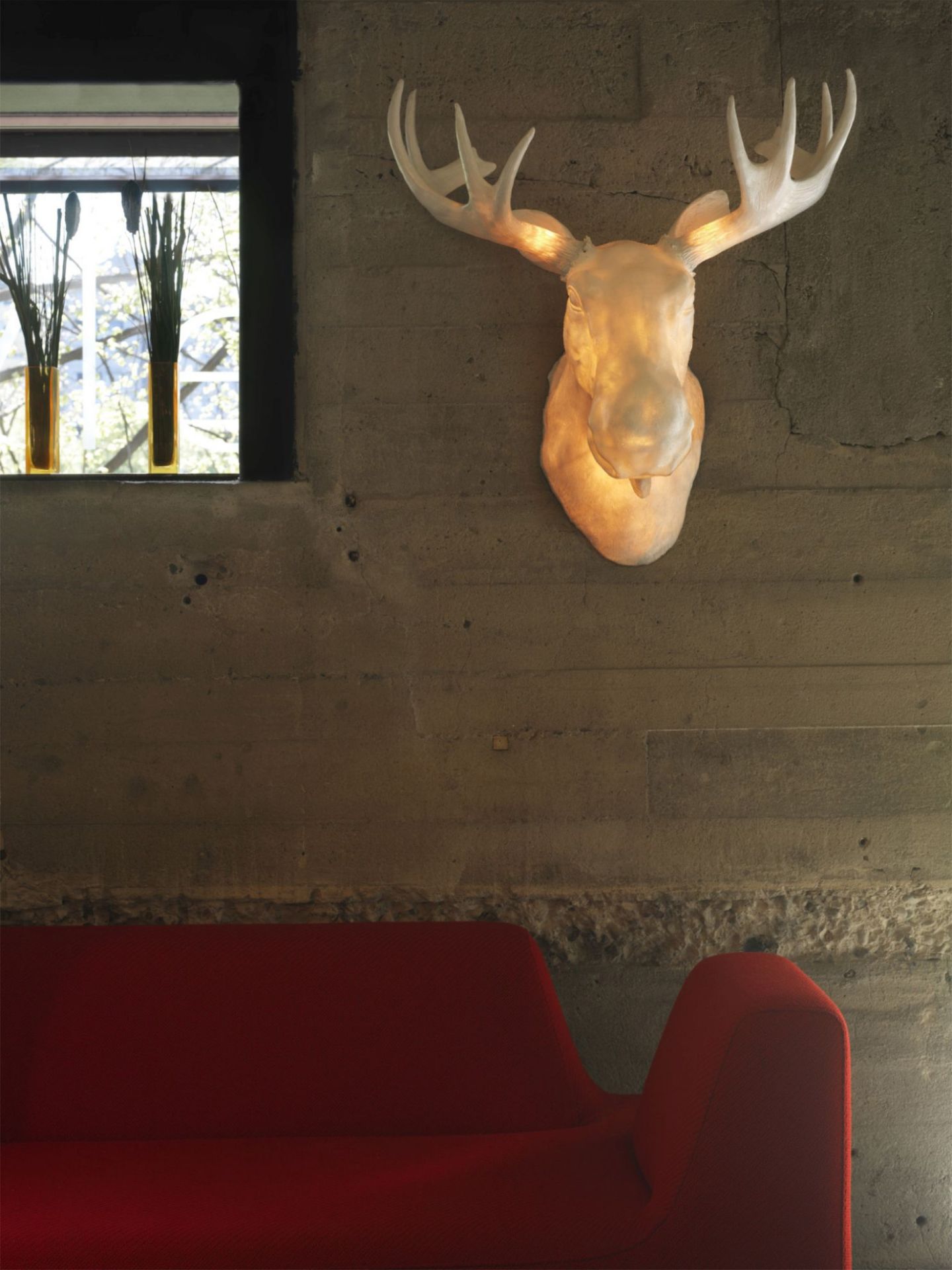 Moo Wandleuchte Northern, Elchkopf-Design, an Betonwand über rotem Sofa. Design Lampe für Wohnzimmer.
