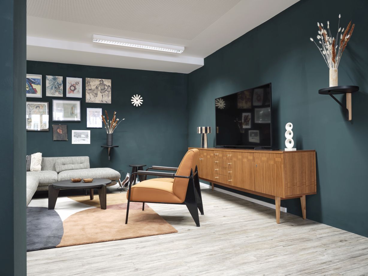 Wohnzimmer mit grauem Vitra Sofa, braunem Sessel und Holzkommode vor dunkelgrüner Wand.