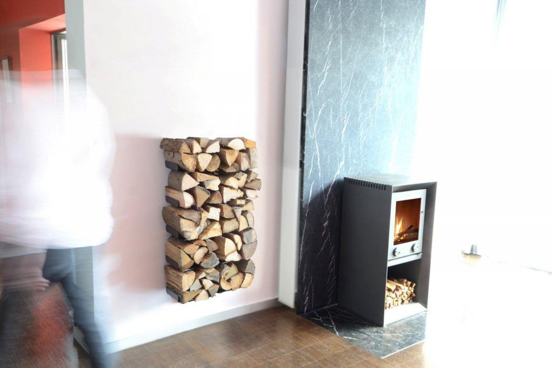 Wooden Tree Kaminholzregal Radius an weißer Wand, daneben Kaminofen mit Holz, modernes Wohnambiente.