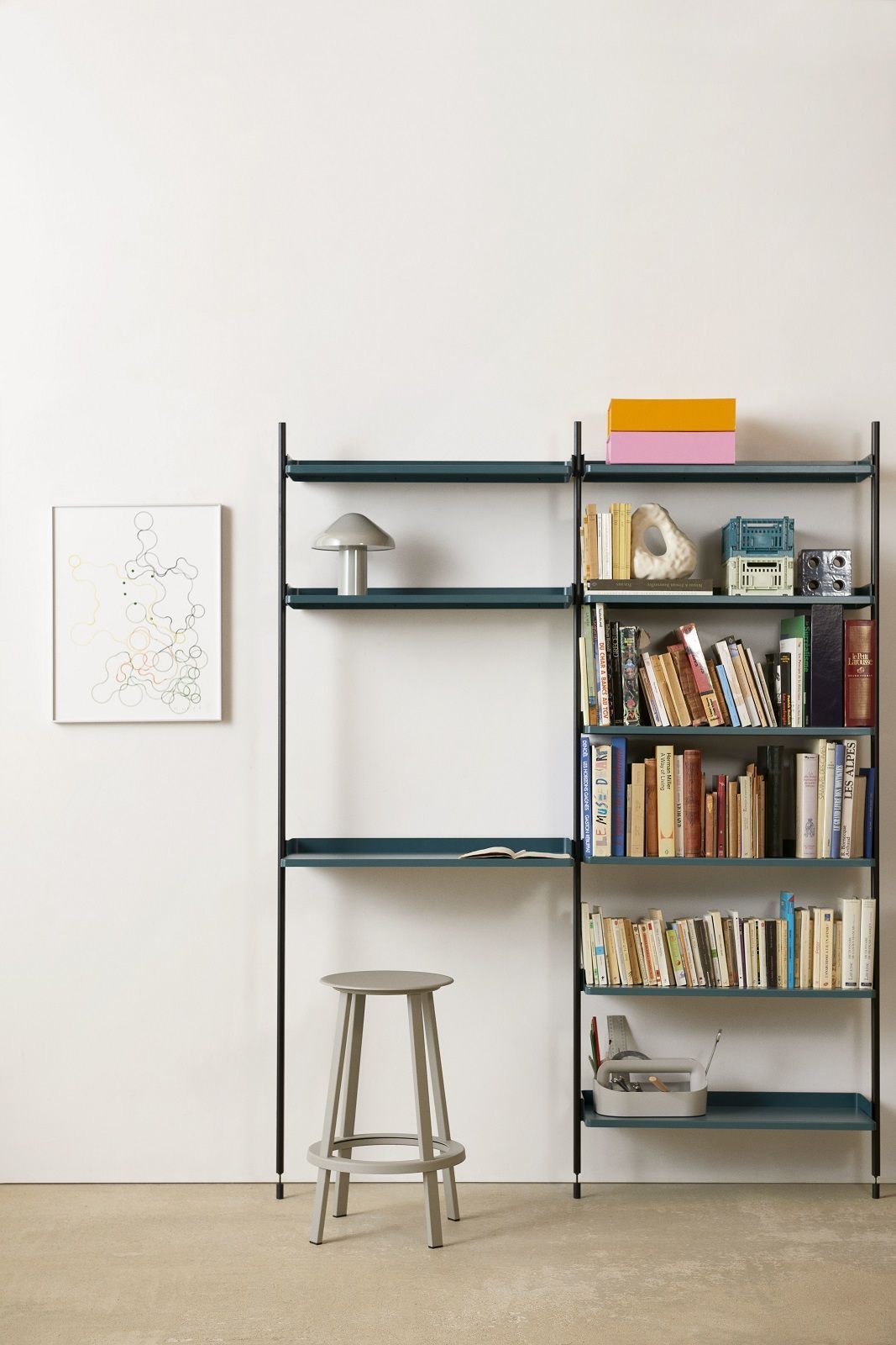Hay Pier System Regal mit zwei Säulen, Bücher, Lampe und Hocker vor einer weißen Wand.